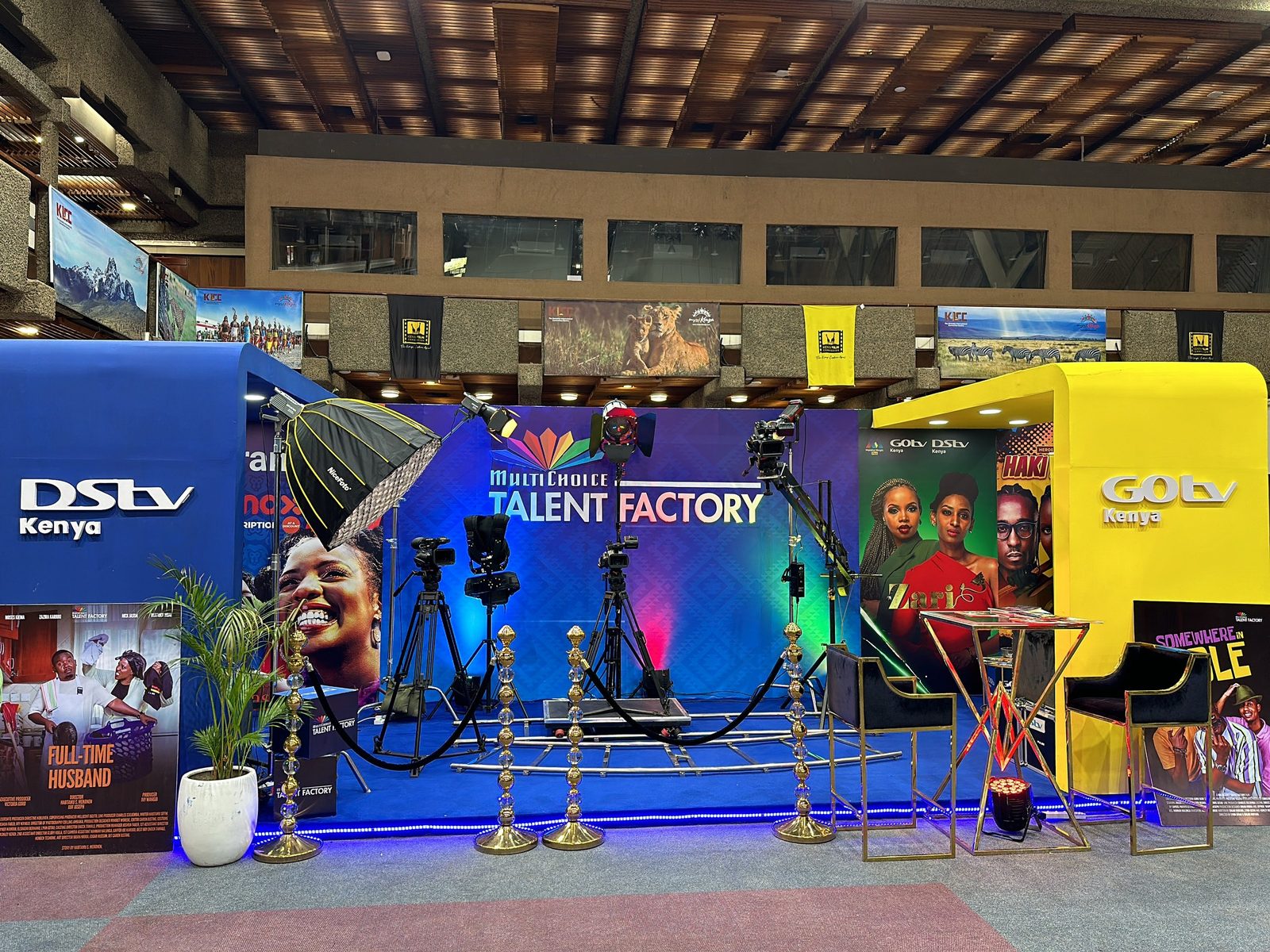 A broadcaster talent-factory stand built under a timber roof, lit in blue with rope-and-post stanchions on a red carpet