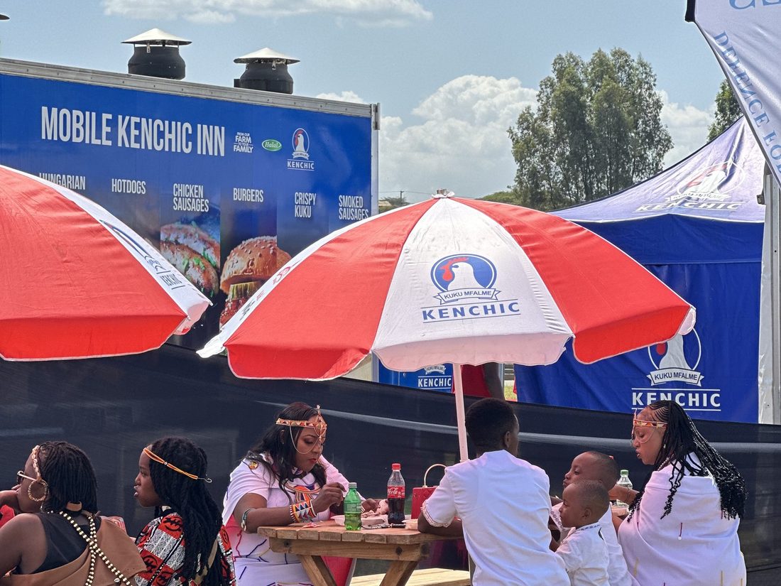 Consumers seated at wooden benches under branded Kenchic umbrellas on a lawn