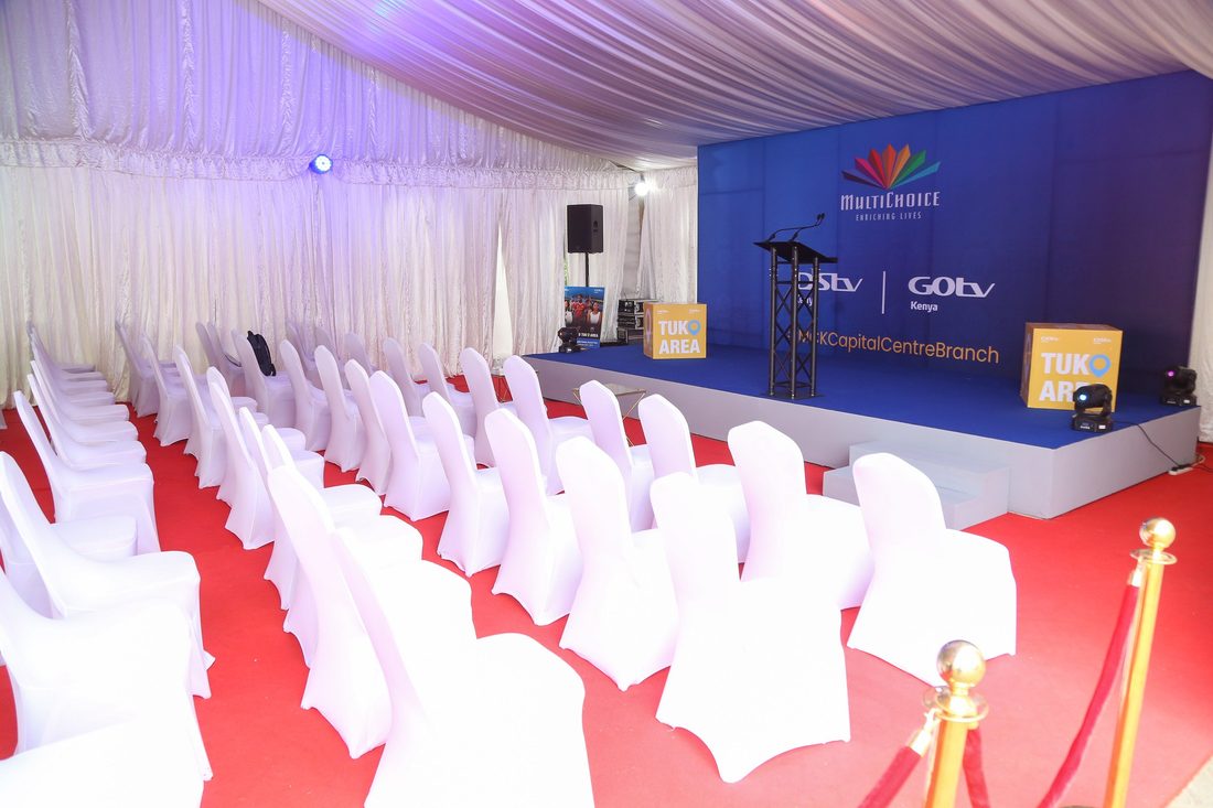 A marquee laid out with a red carpet aisle and covered chairs in front of a branded stage