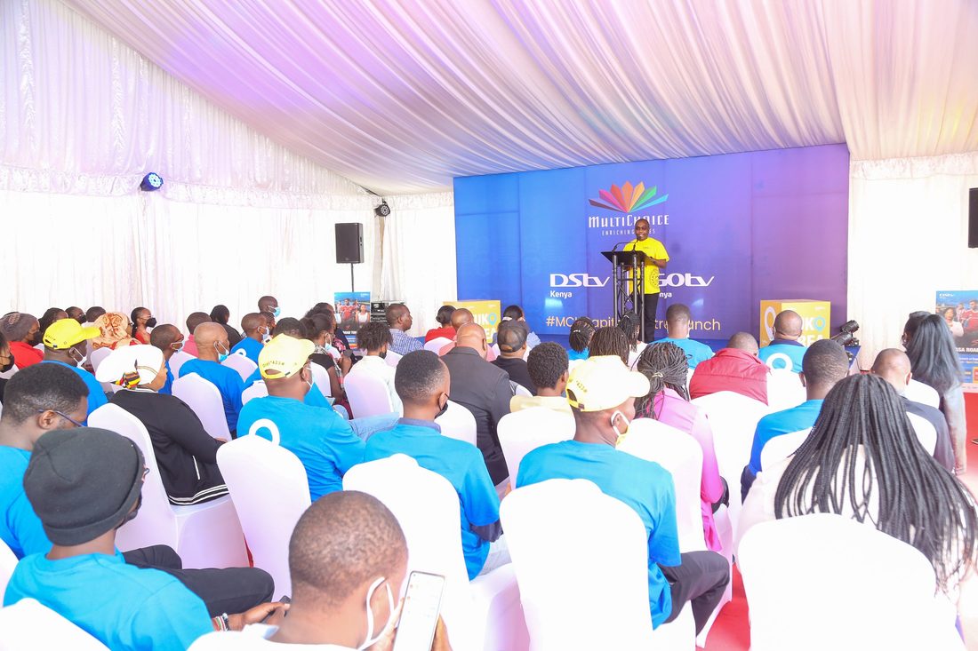 A full audience in branded shirts seated under a marquee facing a lit stage