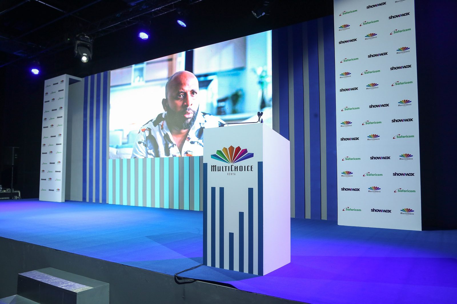 A speaker at a lectern on a lit stage in front of a large screen and a printed wall