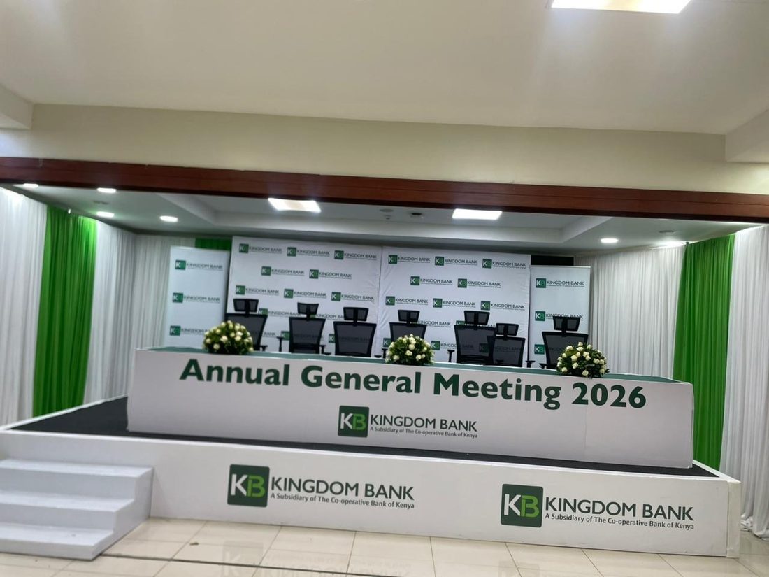 An AGM stage set with tiered white desks and a branded backdrop