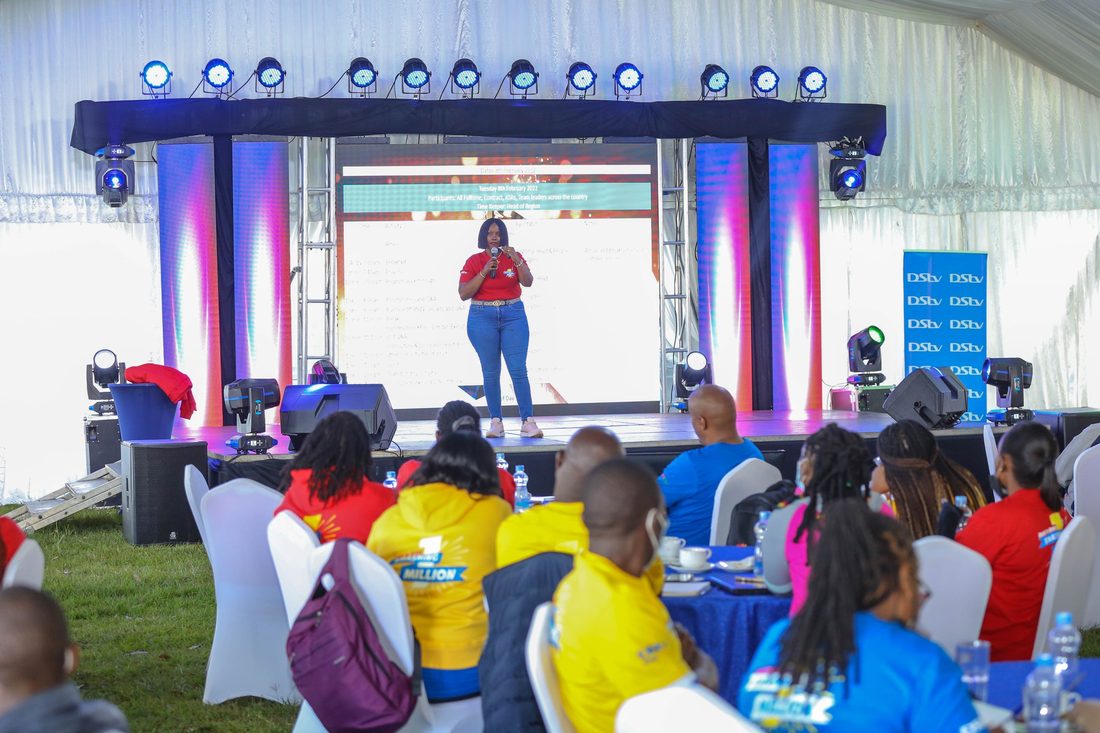 A presenter on a lit marquee stage with delegates watching