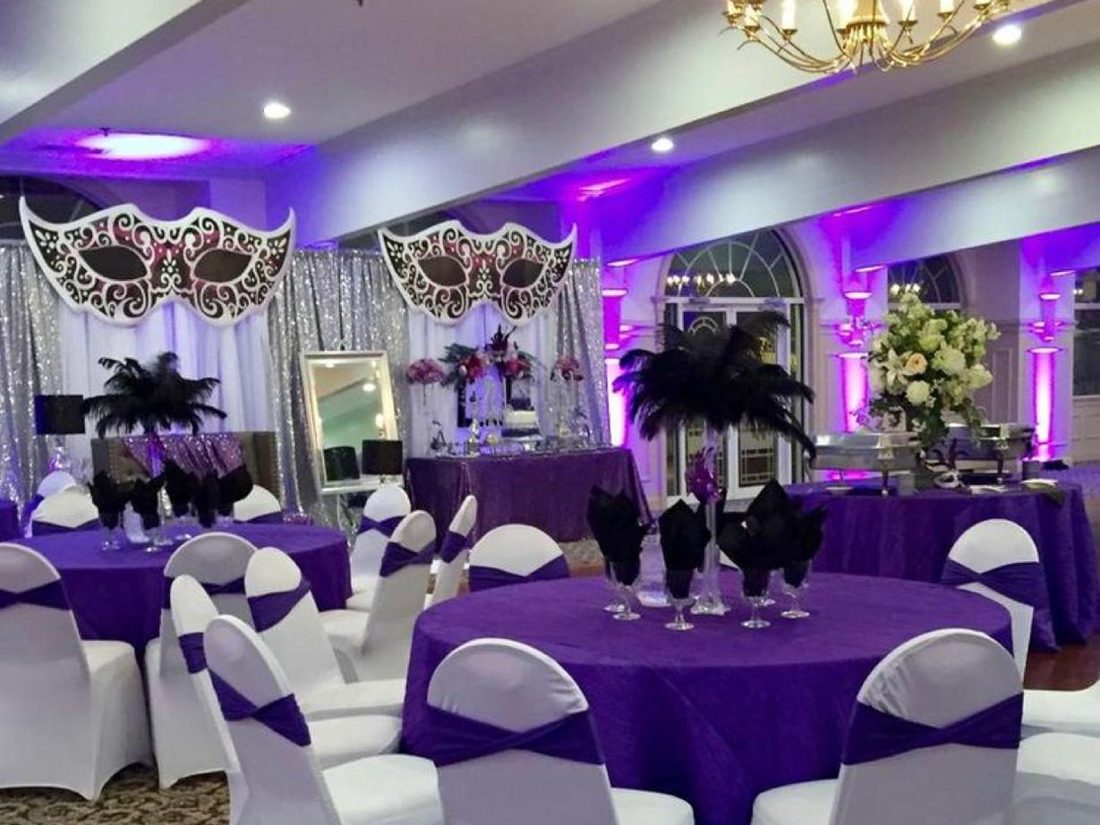 A banquet room under purple uplighting with crystal candelabra and feather masks laid on the tables