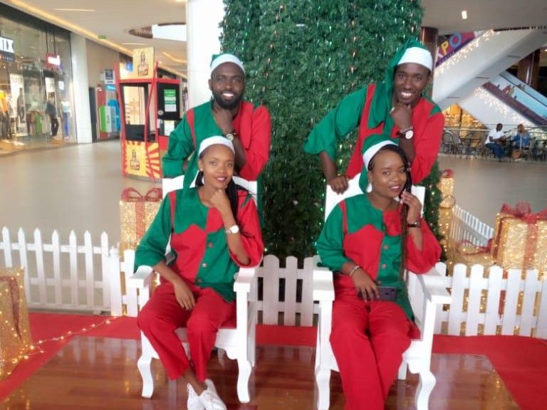 Four costumed performers seated at the base of a shopping-centre Christmas tree