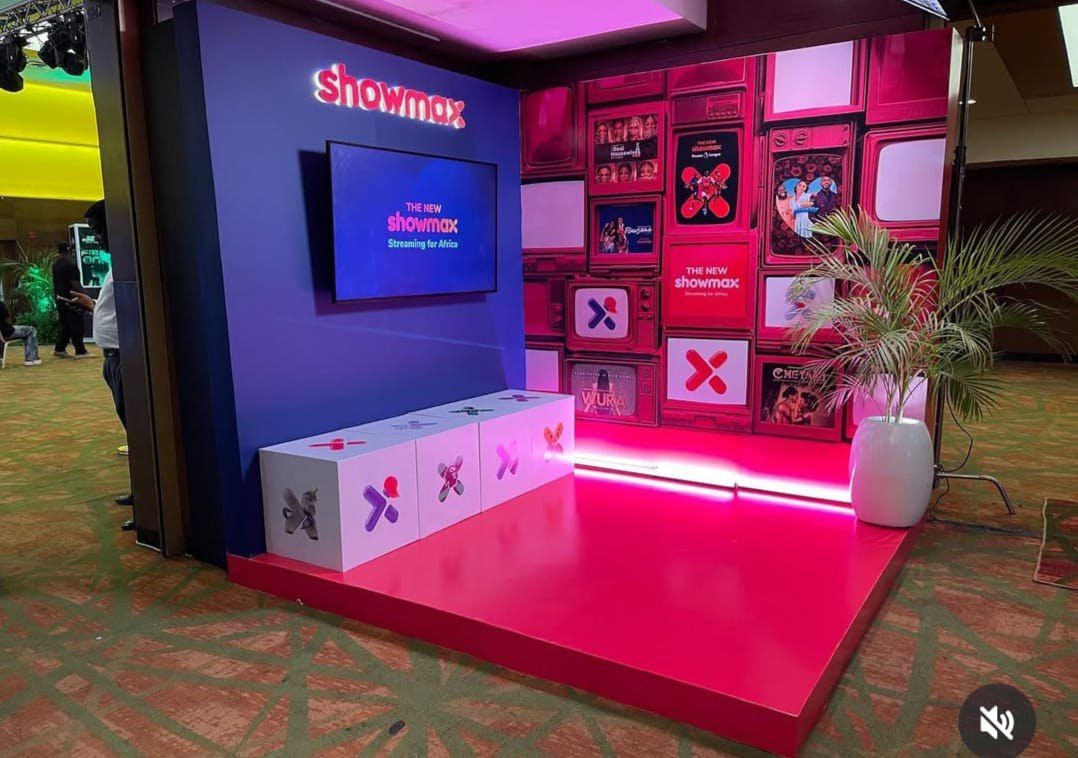 A Showmax stand built around a sculptural X, lit in magenta with a wall of framed title cards behind it