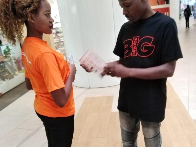 An ambassador in an orange tee handing a leaflet to a consumer who is reading it