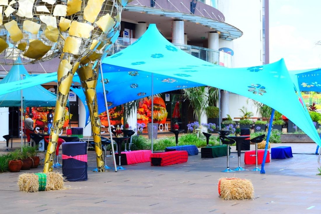 A plaza chill zone with cyan tents, a sculpture and shuka-covered hay bales