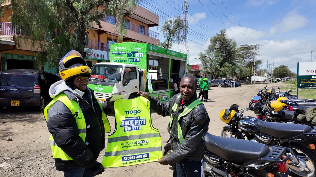 A field representative and a boda rider holding up a branded vest, with a line of motorbikes behind