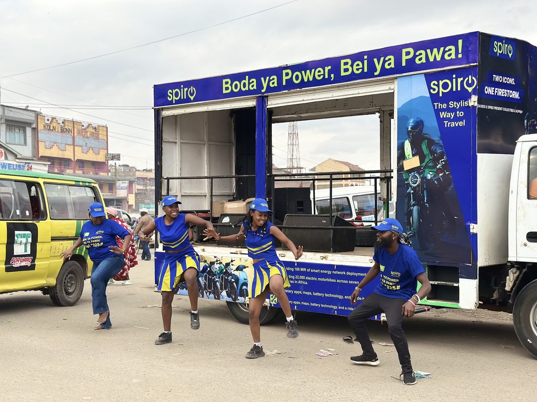 Dancers mid-routine in front of a Spiro Boda ya Power truck on a busy street with matatus passing