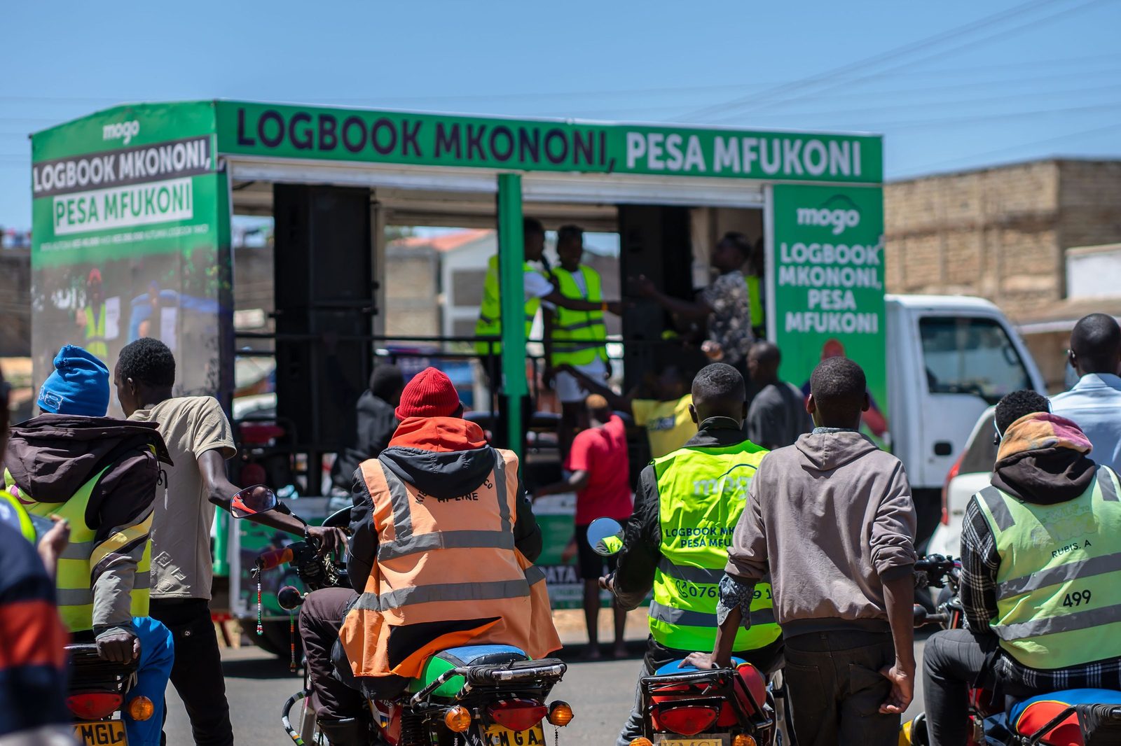 A green branded route truck open at the side with a crowd of motorcycle riders in hi-vis vests gathered around it on a street
