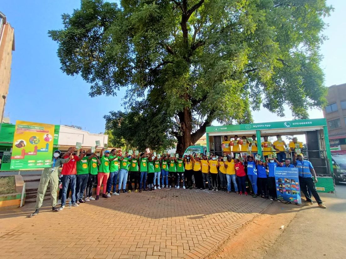 A large team lined up under trees in front of printed product banners