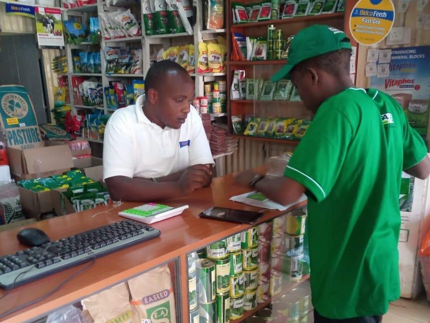 A field representative in a green cap writing an order at an agrovet counter, shelves of animal feed and seed behind