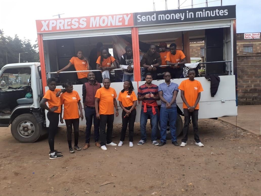An Xpress Money branded truck stage with the field team in front of a row of shops