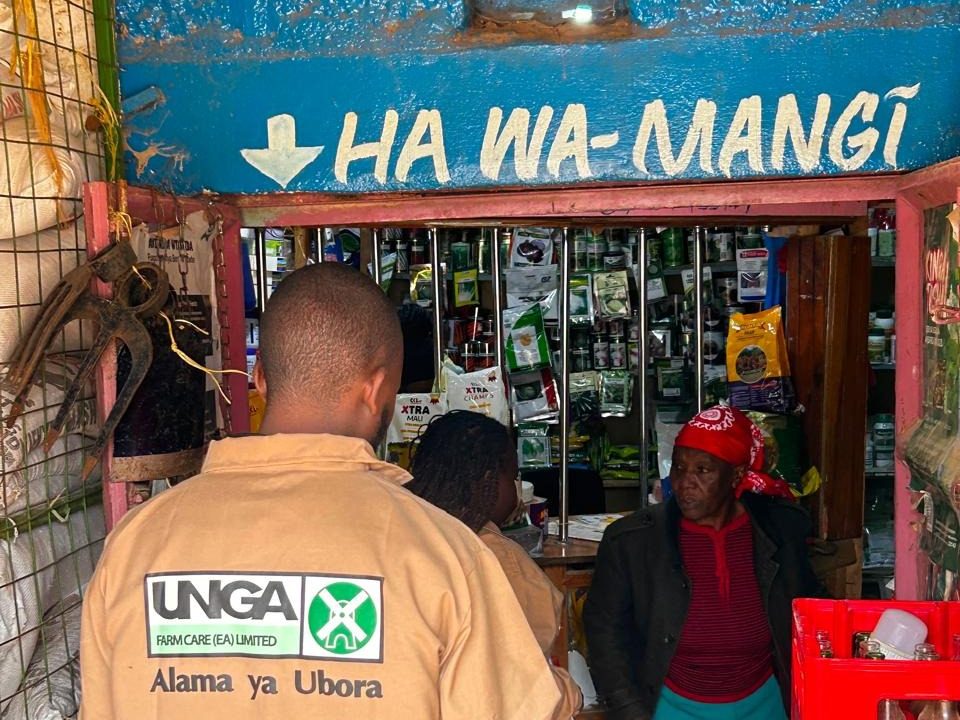 An Unga field representative in branded overalls outside a stocked neighbourhood duka